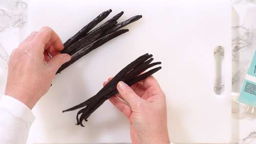 Hands holding Gourmet Grade-A Ugandan Vanilla Beans on a cutting board, showcasing their rich color and quality.